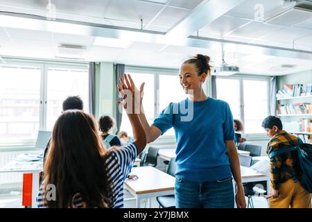 Glückliche Lehrerin, die Schülerinnen High-Five gibt, während sie in der Grundschule im Klassenzimmer steht Stockfoto