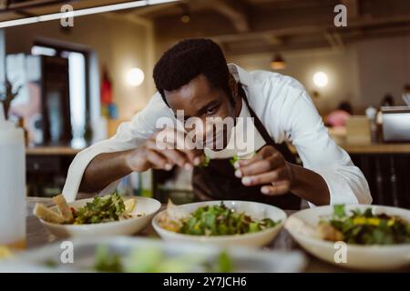 Konzentrierter männlicher Koch garniert Salat mit Koriander, während er sich in der kommerziellen Küche beugt Stockfoto