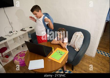 Zwei Kinder bereiten sich auf die Schule vor. Ein Junge packt seinen Rucksack, während ein Mädchen auf einen Laptop schaut. Schulsachen und Rucksäcke liegen auf dem Tisch. Stockfoto