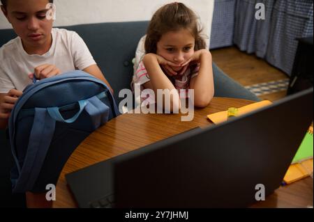 Zwei Kinder, die zu Hause mit einem Laptop und Schulmaterialien auf einem Holztisch pädagogisch tätig sind. Stockfoto