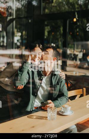 Junge Paare teilen sich zärtliche Momente in einem Café, umgeben von Grün und natürlichem Licht, während sie sich bei warmen Getränken und verspielten Gesten verbinden Stockfoto