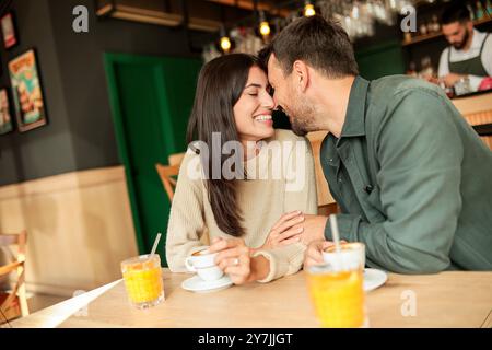 Ein Paar teilt sich einen freudigen Moment in einem stilvollen Café, schlürft Kaffee und genießt hellen Orangensaft, während es gemeinsam lacht, umgeben von einem einladenden Restaurant Stockfoto