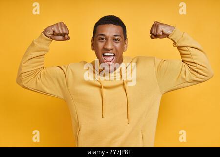 Ein verspielter, gemischter Mann zeigt seinen Bizeps, während er vor gelbem Hintergrund steht Stockfoto
