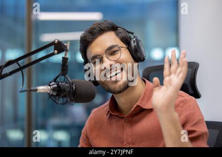 Ein lächelnder Mann im Studio spricht mit Kopfhörern ins Mikrofon und erstellt Podcasts. Ein freundliches Wehen in Richtung Kamera deutet auf eine ansprechende Interaktion hin. Stockfoto