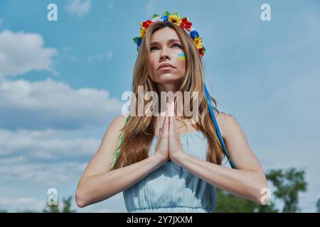 Schöne junge ukrainische Frau in Blumenkrone betend mit blauem Himmel im Hintergrund Stockfoto