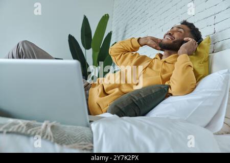 Entspannter junger afrikanischer Mann im Kopfhörer, der Musik genießt, während er zu Hause im Bett liegt Stockfoto