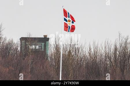 Kirkenes, Norwegen, 16. Oktober 2023: Die norwegische Flagge weht im kalten Wind, dahinter ein russischer Beobachtungsposten. (Foto: Andreas Haas/dieBildmanufak Stockfoto