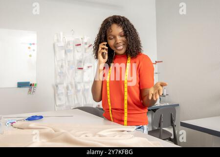Schneiderin in orangefarbenem T-Shirt, der am Telefon spricht, während er Stoff schneidet Stockfoto