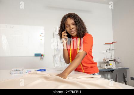 Schneiderin in orangefarbenem T-Shirt, der am Telefon spricht, während er Stoff schneidet Stockfoto