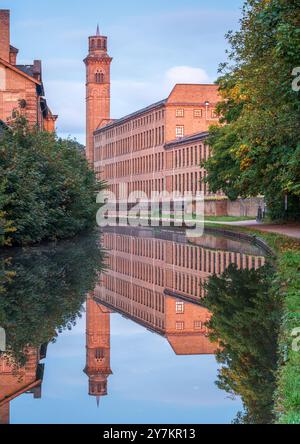 Das New Mill-Gebäude in Salts Mill, Saltaire, leuchtet an einem ruhigen Septembermorgen im reflektierten Licht eines lebhaften Sonnenaufgangs in Yorkshire in einem tiefen Orange. Stockfoto