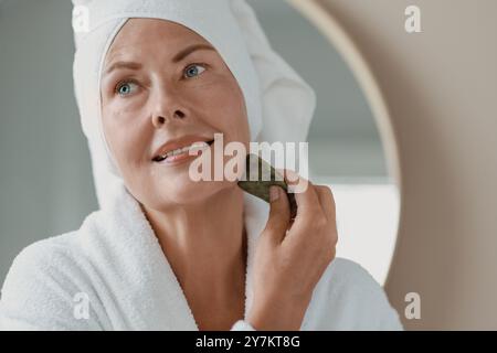 Schöne junge Frau, die eine Gesichtsmassage mit Gua-Sha-Werkzeug drinnen macht Stockfoto