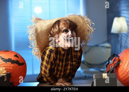 Süßer Junge, der wie Vogelscheuche gekleidet ist, mit festlichem Dekor und Geschenkboxen drinnen in der Nacht. Halloween-Feier Stockfoto