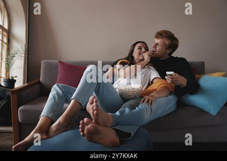 Glückliches junges Paar, das Fernsehen sieht und Popcorn isst, während es sich auf der Couch zu Hause entspannt Stockfoto