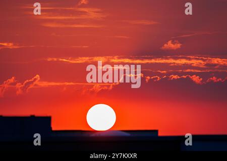 Atemberaubende feurige Sonnenuntergangsszene mit leuchtendem rotem Himmel, glühender Sonne, die hinter städtischen Silhouetten untergeht und eine warme dramatische Atmosphäre schafft Stockfoto