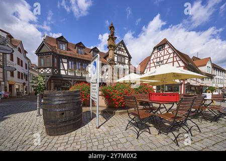 Marktbrunnen mit Geranien, Tischen, Stühlen und Sonnenschirmen eines Restaurants und Fachwerkhäusern unter blauem Himmel mit Kumuluswolken auf der Marke Stockfoto