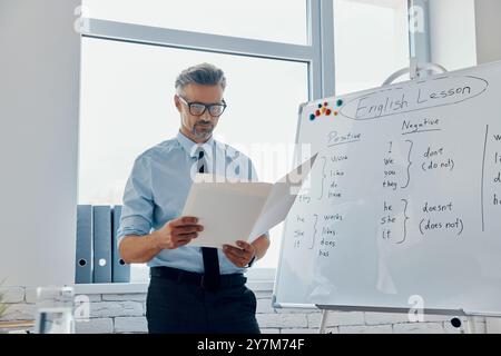 Selbstbewusster Englischlehrer, der die Unterlagen durchgeht, während er im Klassenzimmer neben dem Whiteboard steht Stockfoto