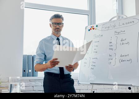 Ein selbstbewusster Englischlehrer hält Papiere, während er im Klassenzimmer neben dem Whiteboard steht Stockfoto