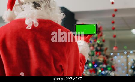 Angestellter in festlich verzierten Einkaufszentren im Weihnachtsmann-Anzug, der ein grünes Bildschirmtelefon hält, eine Pause von der Arbeitsschicht einlegt und Videos auf Chroma Key Screen ansieht, Nahaufnahme Stockfoto