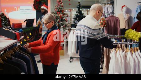 Ältere Frau und Ehemann stöbern durch Kleiderständer im weihnachtlich dekorierten Bekleidungsgeschäft. Älteres Paar glücklich, nachdem es perfekte Blazer gefunden hat, die man dem Enkel schenken kann Stockfoto