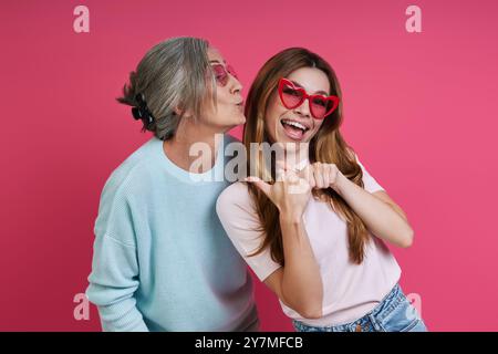 Verspielte Mutter und Erwachsene Tochter in flippiger Brille, die Spaß vor rosa Hintergrund haben Stockfoto