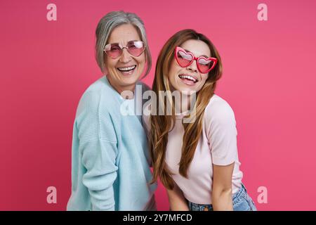 Glückliche Mutter und erwachsene Tochter tragen eine funkige Brille, während sie zusammen vor rosa Hintergrund stehen Stockfoto