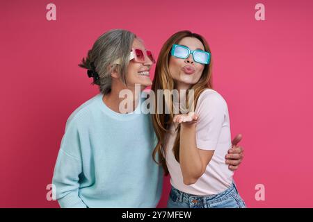 Verspielte Mutter und Erwachsene Tochter in flippiger Brille, die Spaß vor rosa Hintergrund haben Stockfoto