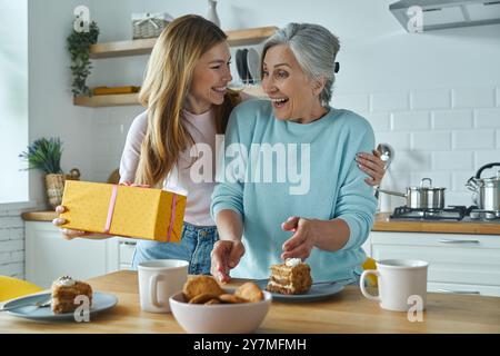 Überraschte Mutter, die eine Geschenkbox von ihrer Tochter bekam, während beide in der Küche saßen Stockfoto
