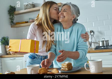 Junge Frau, die ihre Mutter küsst und eine Geschenkbox hält, während sie Zeit in der Küche verbringt Stockfoto