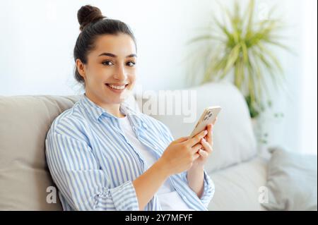 Fröhliche, glückliche, schöne inder- oder arabianerin in lässiger Kleidung, die auf einer bequemen Couch sitzt und online mit Freunden SMS per Smartphone sendet, Online-Shops durchstöbert, die Kamera ansieht und lächelt Stockfoto