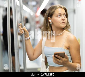 Ein junges Mädchen, das in einem U-Bahn-Wagen steht, das Telefon und kabellose Kopfhörer in der Luft hält Stockfoto