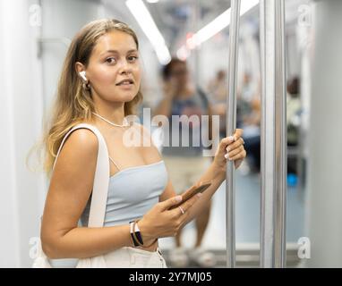 Positives, junges Mädchen, das in einem U-Bahn-Wagen steht und Telefon und kabellose Kopfhörer auf Sendung hält Stockfoto