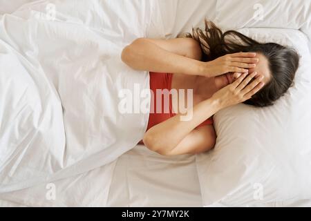 Draufsicht einer depressiven jungen Frau, die im Bett liegend das Gesicht mit den Händen bedeckt Stockfoto