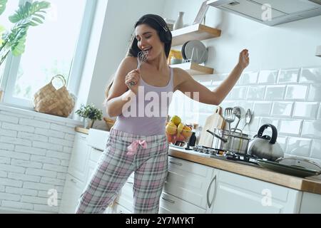 Glückliche junge Frau, die den Schneebesen als Mikrofon benutzt und beim Kochen in der Küche tanzt Stockfoto