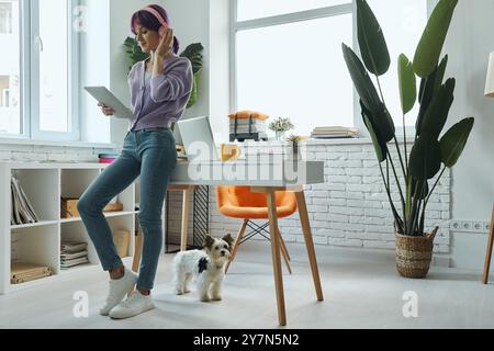 Schöne junge Frau in Kopfhörern, die ein digitales Tablet benutzt, während sie in der Nähe ihres Arbeitsplatzes steht Stockfoto