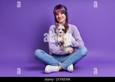 Schöne junge Frau, die einen kleinen Hund trägt und lächelt, während sie vor violettem Hintergrund sitzt Stockfoto