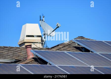 Solarpaneele auf einem braunen Schindeldach mit verschwommenem Kamin und Satellitenschüssel im Hintergrund. Selektiver Fokus Stockfoto