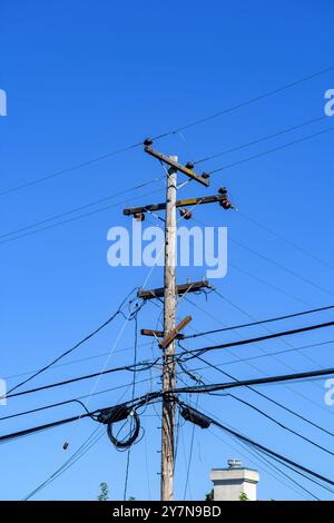 Ein hoher hölzerner Versorgungsmast mit mehreren Drähten und Kabeln, die in verschiedene Richtungen unter klarem blauem Himmel verlaufen. Der Stab und die Kabel sind verwittert Stockfoto