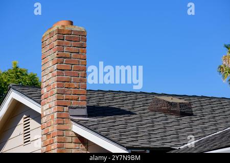 Ein Wohndach mit einem roten Ziegelkamin, dessen Schornsteinkappe auf den schwarzen Schindeln verlagert liegt. Stockfoto