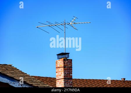 Eine traditionelle Dachantenne auf einem gemauerten Kamin erhebt sich über dem braunen Schindeldach eines Hauses Stockfoto