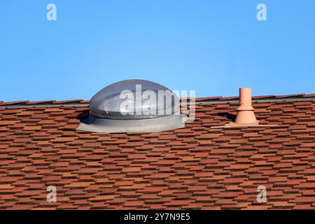 Ein Wohndach mit roten Asphaltschindeln mit kuppelförmigem Oberlicht und einem kleinen Entlüftungsrohr. Nahaufnahme. Stockfoto