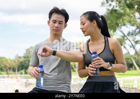 Ein gesundes, fittes asiatisches Paar oder Freunde ruhen sich aus, nachdem sie gemeinsam in einem grünen Park gelaufen sind, Wasser trinken und ihre Laufmeilen mit einem Smartw überprüfen Stockfoto