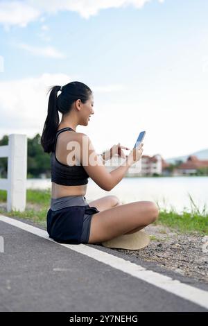 Ein wunderschönes, sportliches asiatisches Mädchen in Sportbekleidung sitzt am See in einem Park und nutzt ihr Smartphone, um sich nach dem Laufen auszuruhen. Stockfoto