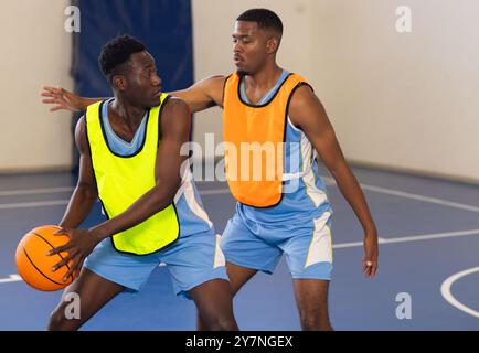 Zwei afroamerikanische Männer in Sportrikots spielen Basketball und treten auf dem Hallenplatz an Stockfoto
