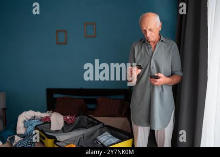 Buchungs-App: Pensionierter Tourist verwendet Smartphone, während er zu Hause sein Gepäck packt und sich auf den Urlaub vorbereitet Stockfoto