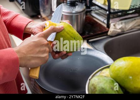 Frau schält reife und saftige Mangobrüchte mit einem scharfen Messer in der Küche Stockfoto