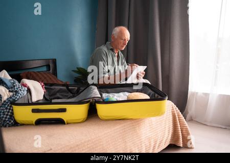 Urlaubs-, Urlaubs- und Reisekonzept. Reisender älterer Mann mit Checkliste nach dem Packen, Vorbereitung der Sachen in das Gepäck für die Reise sitzend Stockfoto