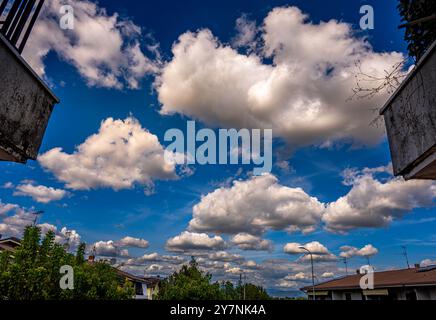 Dieses Bild zeigt einen lebendigen Himmel voller flauschiger weißer Wolken vor einem tiefblauen Hintergrund. Darunter Dächer und grüne Bäume eines Vorstadtgebiets. Stockfoto