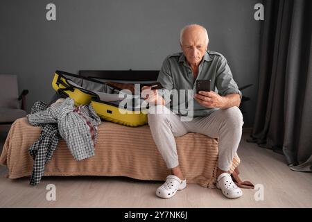 Reisender Mann sitzt mit Koffer und macht Urlaub Tour Reservierung. Großvater benutzt Handy und Bucht das Hotel online mit Kreditkarte und Stockfoto