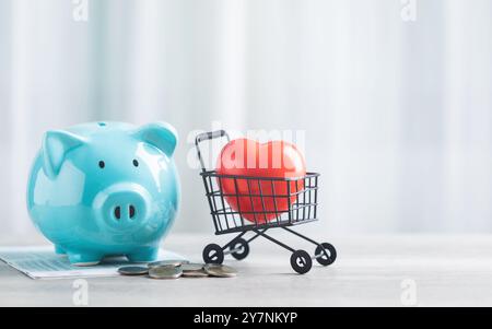 Eine blaue Schweinebank, ein Münzhaufen und ein rotes Herz in einem Einkaufswagen sitzen auf einem Tisch vor einem hellen weißen Hintergrund. Das Bild symbolisiert das Speichern von m Stockfoto
