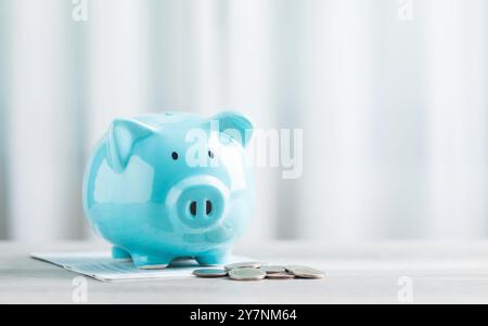 Eine blaue Schweinebank, ein Münzhaufen und ein Bankbuch stehen auf einem Tisch vor einem hellen weißen Hintergrund. Das Bild vermittelt die Idee, Geld zu sparen Stockfoto